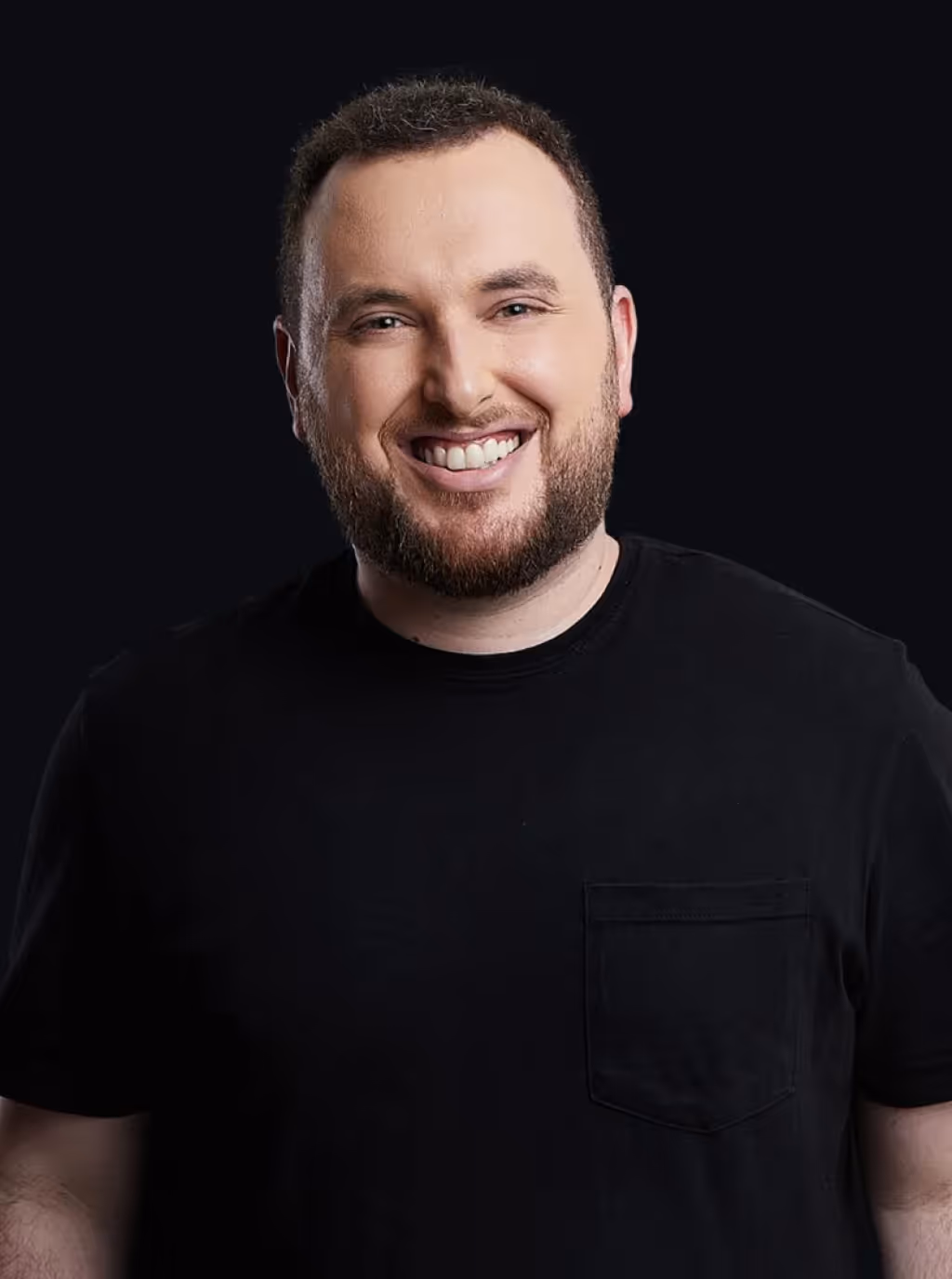 Smiling man with short hair and beard wearing a black t-shirt against a dark background.